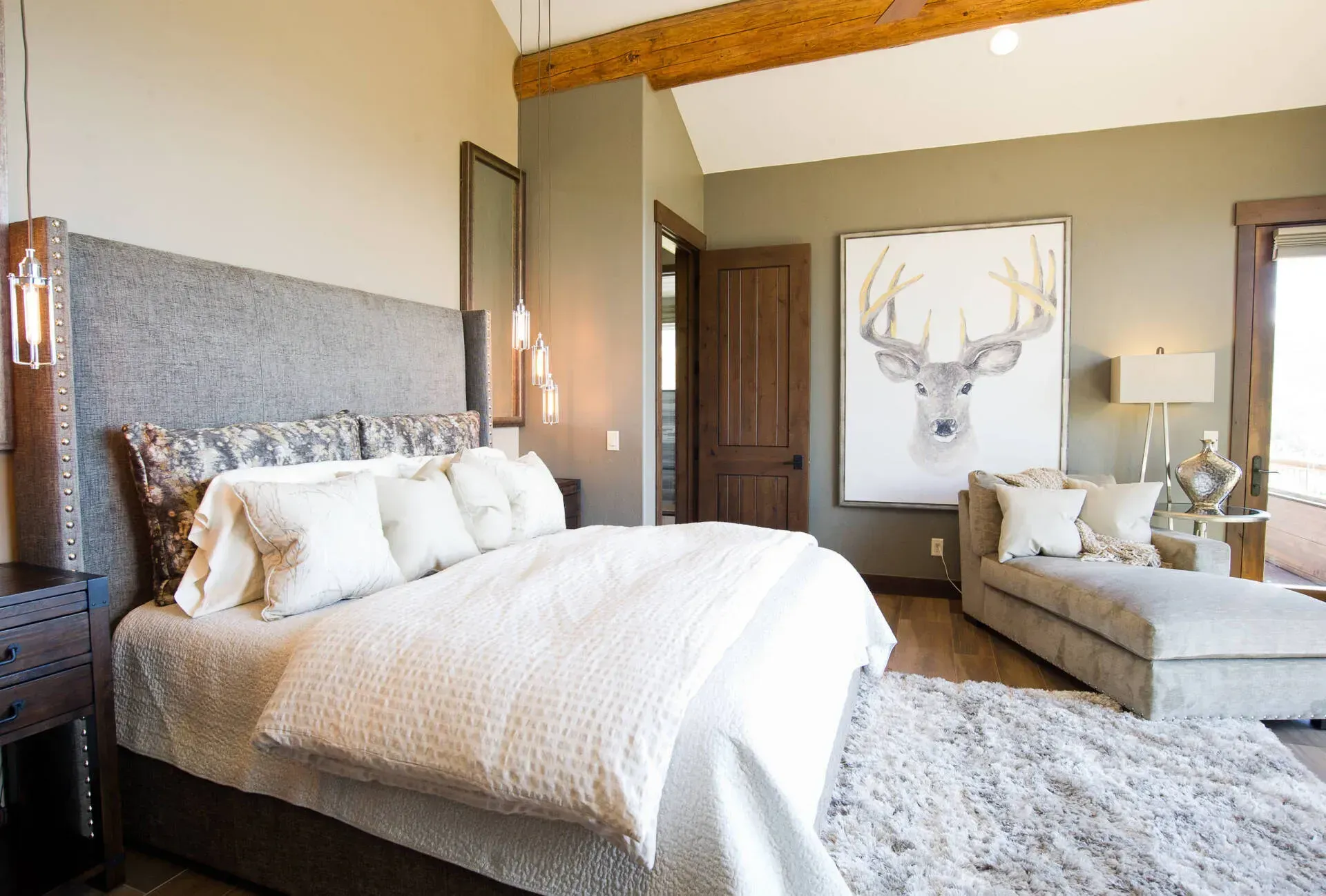 Bedroom with a large bed, chaise lounge, and artwork; neutral tones, wooden beams.