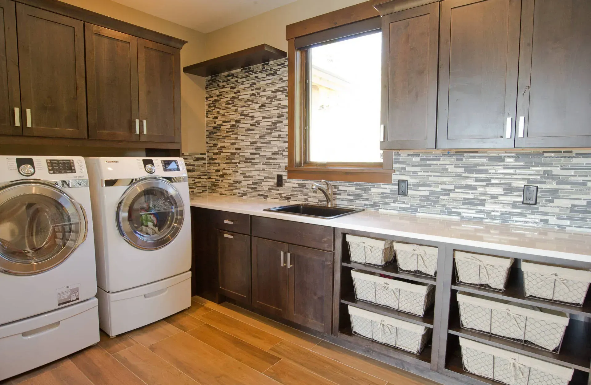 Laundry room with washer, dryer, dark cabinets, a sink, and baskets on shelves; mosaic tile backsplash.