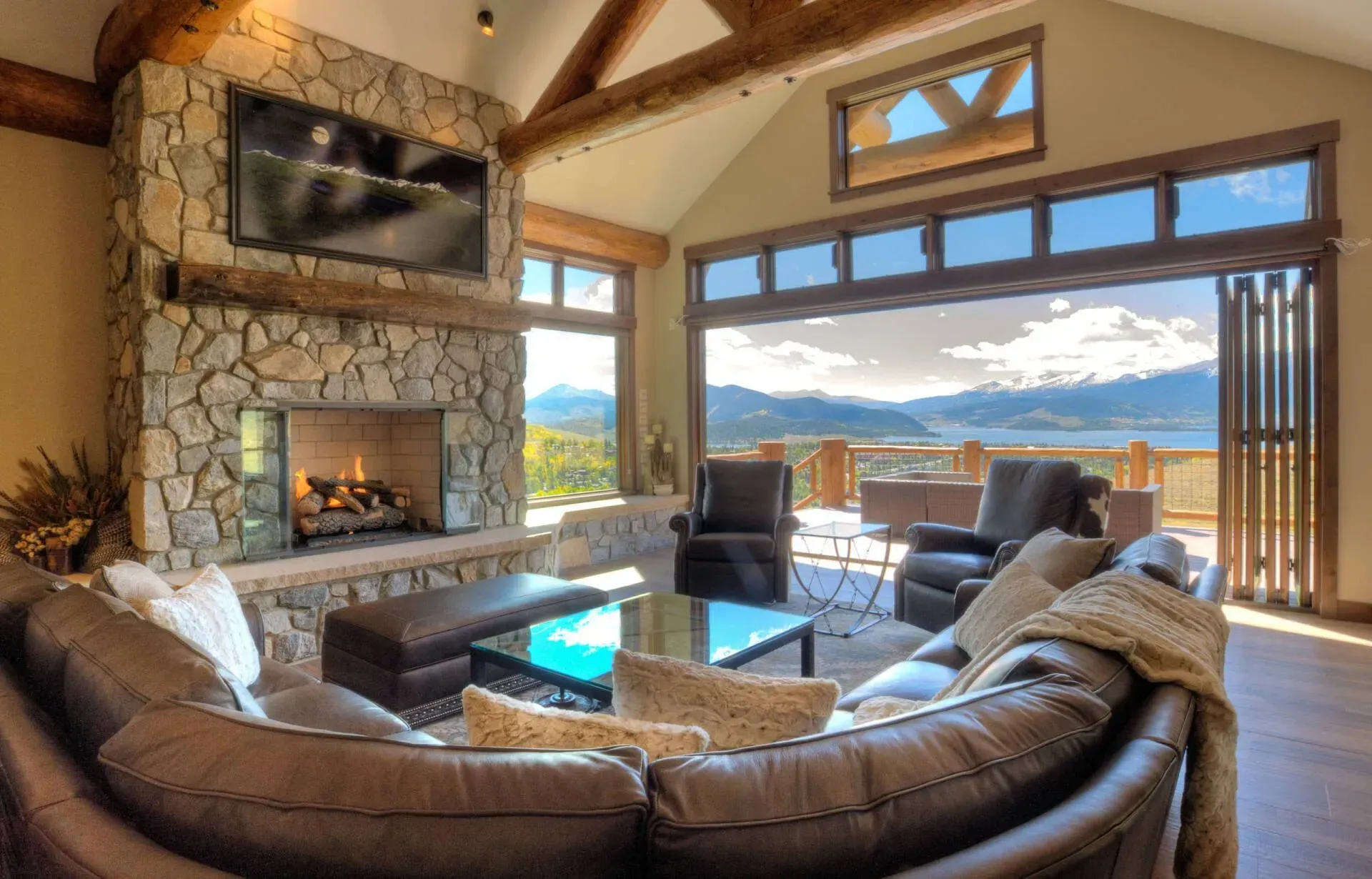Cozy living room with stone fireplace, large sectional sofa, and expansive mountain views through open glass doors.