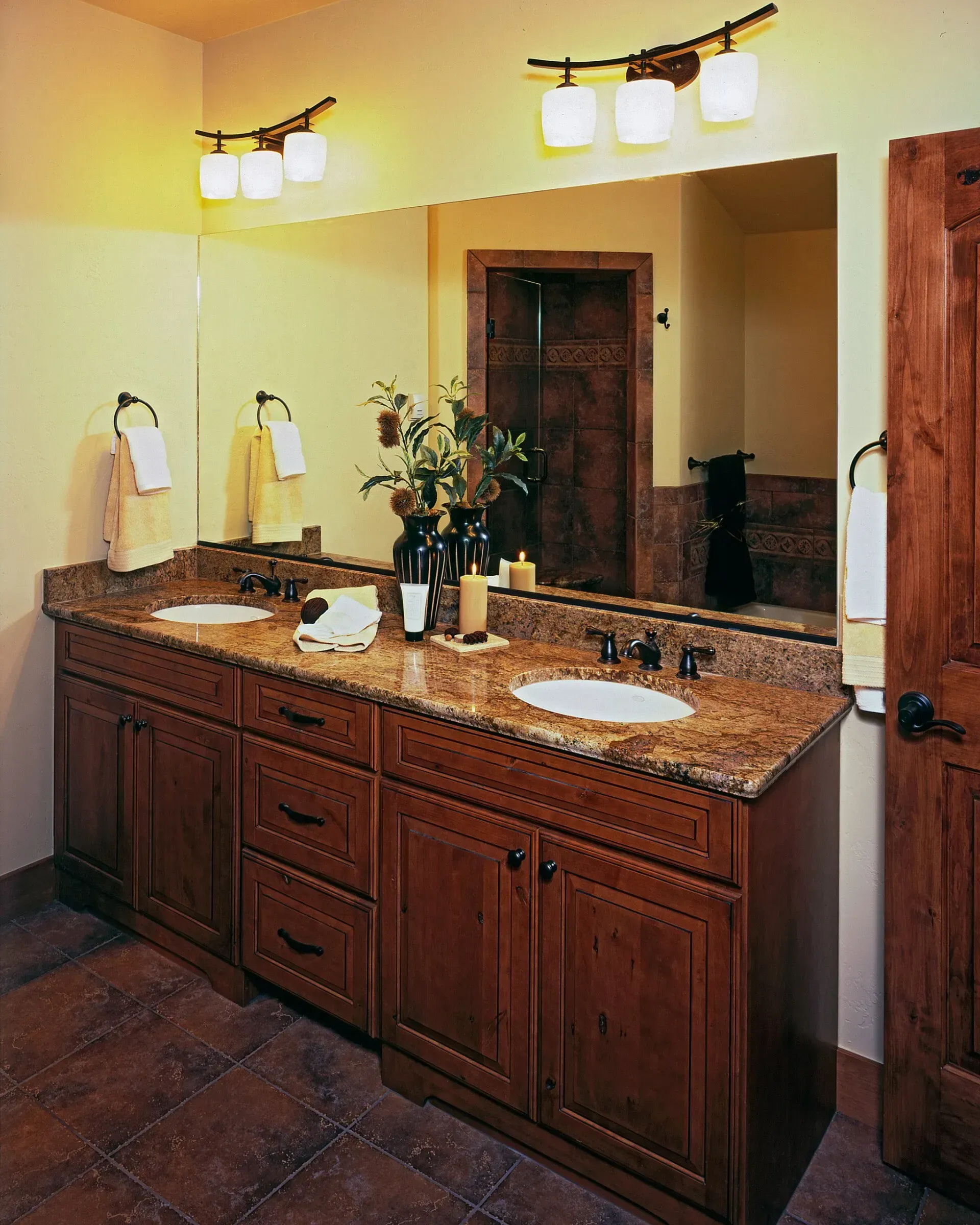 Bathroom with double vanity, granite countertop, brown cabinets, large mirror, and wall sconces.