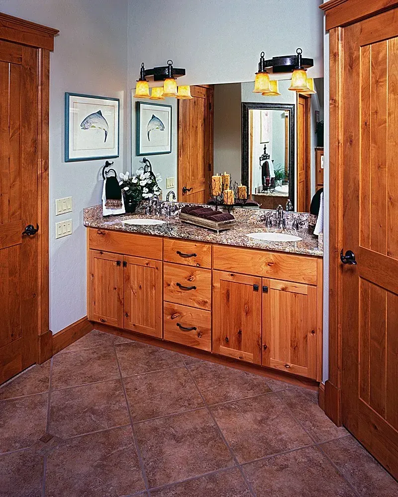 Bathroom with wood cabinets, double sinks, and a large mirror. Brown tile floor, light gray walls, and framed art.