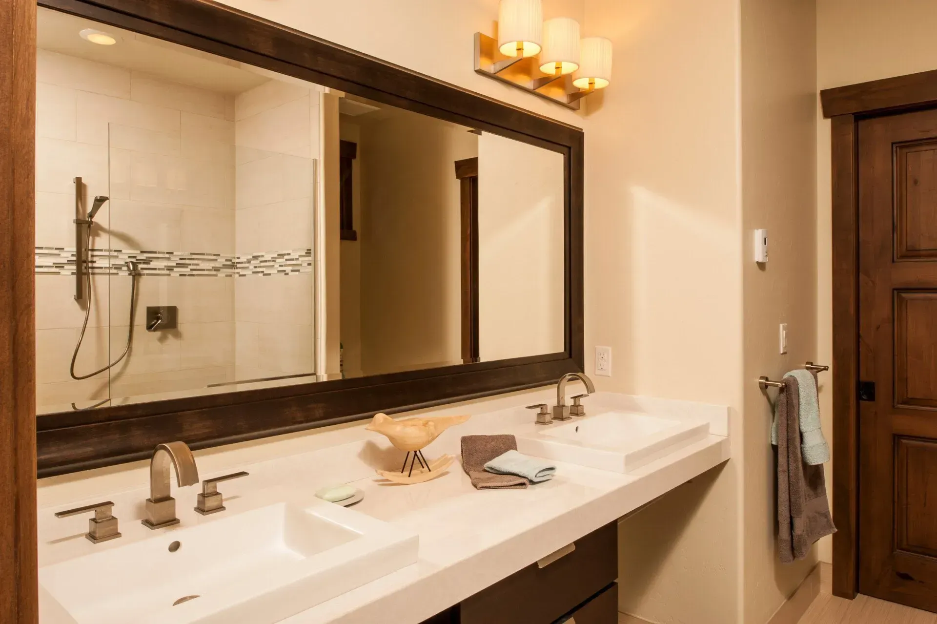 Bathroom with double sinks, large mirror, wooden door, and shower visible.