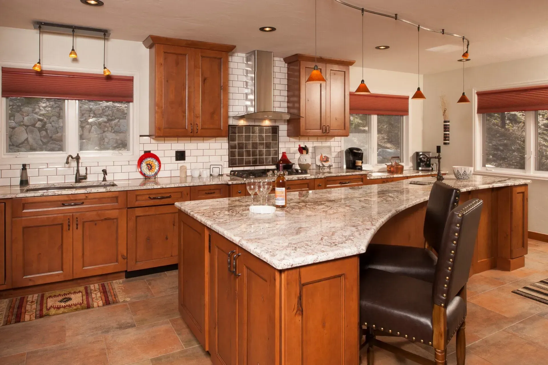 A warm-toned kitchen with a large island, wooden cabinets, granite countertops, and red window shades.