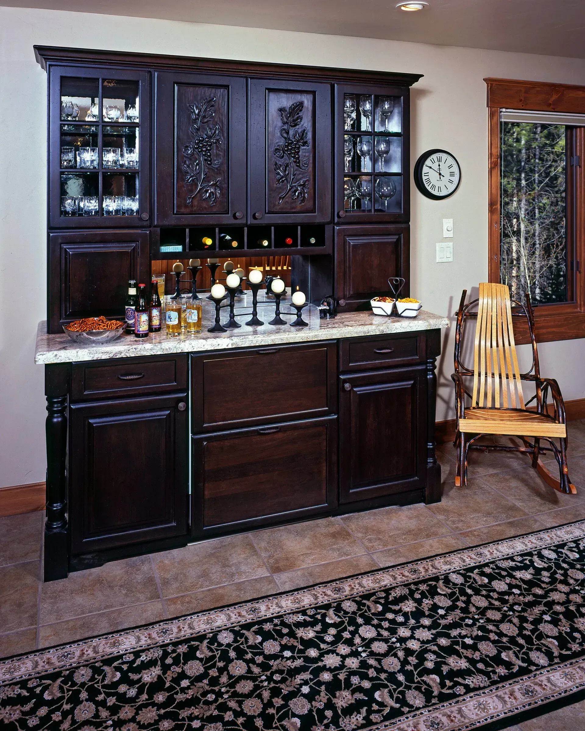 Dark wood bar with granite countertop, cabinet storage, and glass-door display. A rocking chair sits nearby.