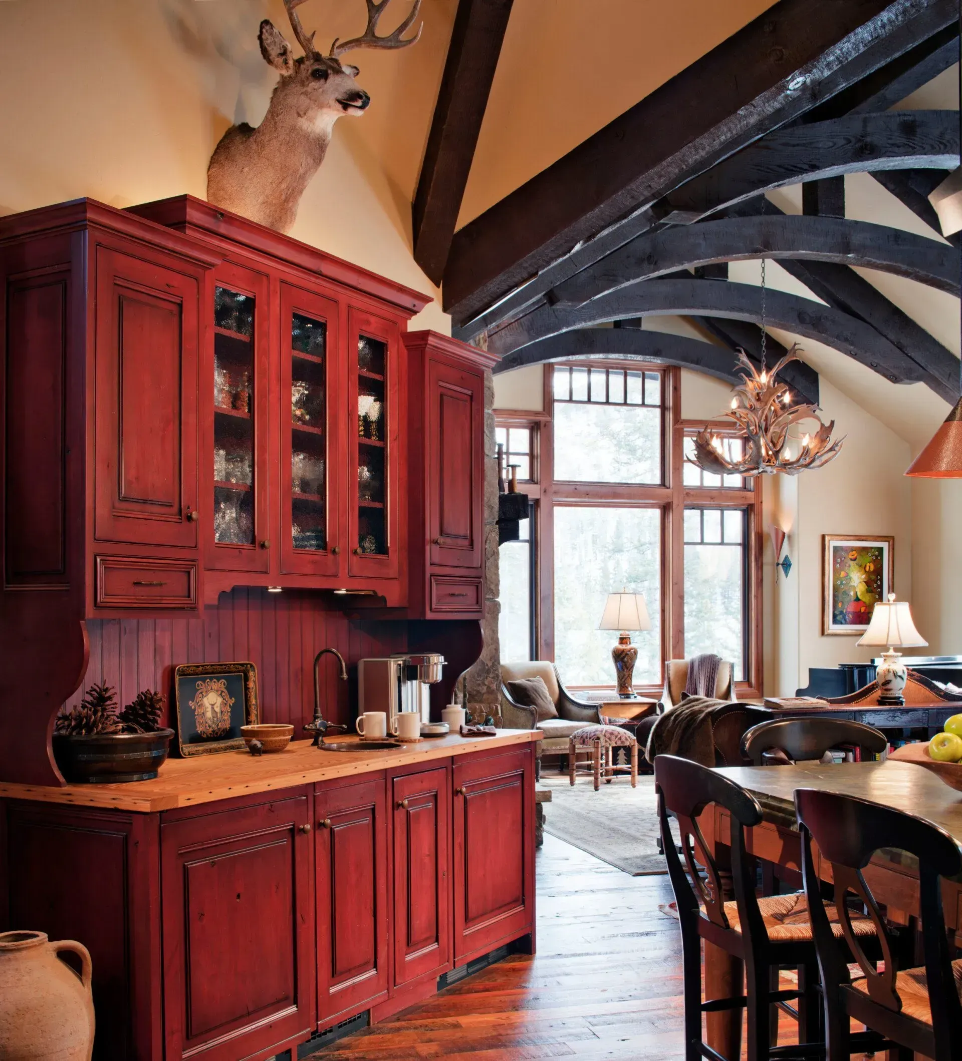 Red kitchen cabinet with deer head above, in a room with a large window and wood beams.
