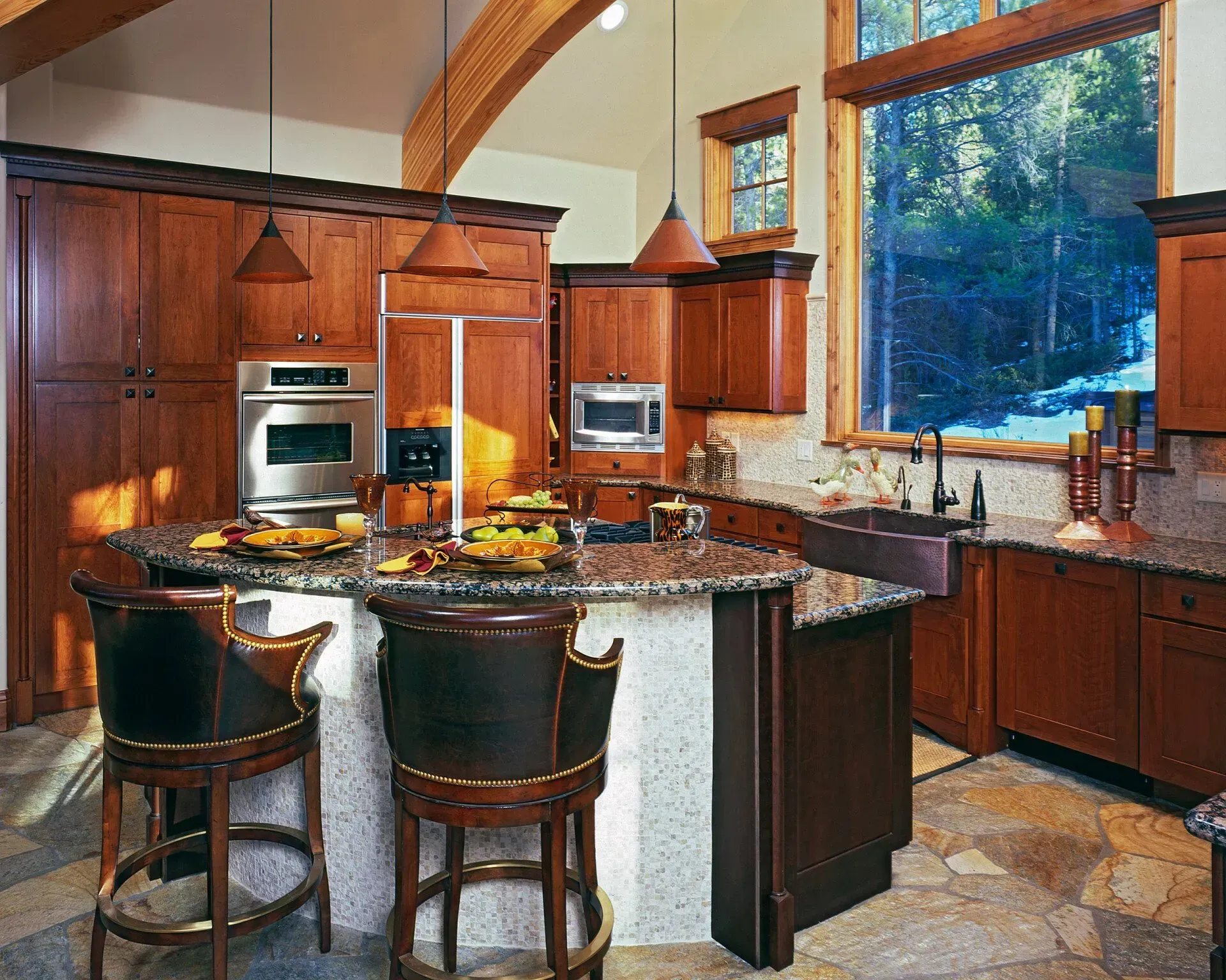 A kitchen with brown cabinets, a granite countertop island, and a large window overlooking a forest.