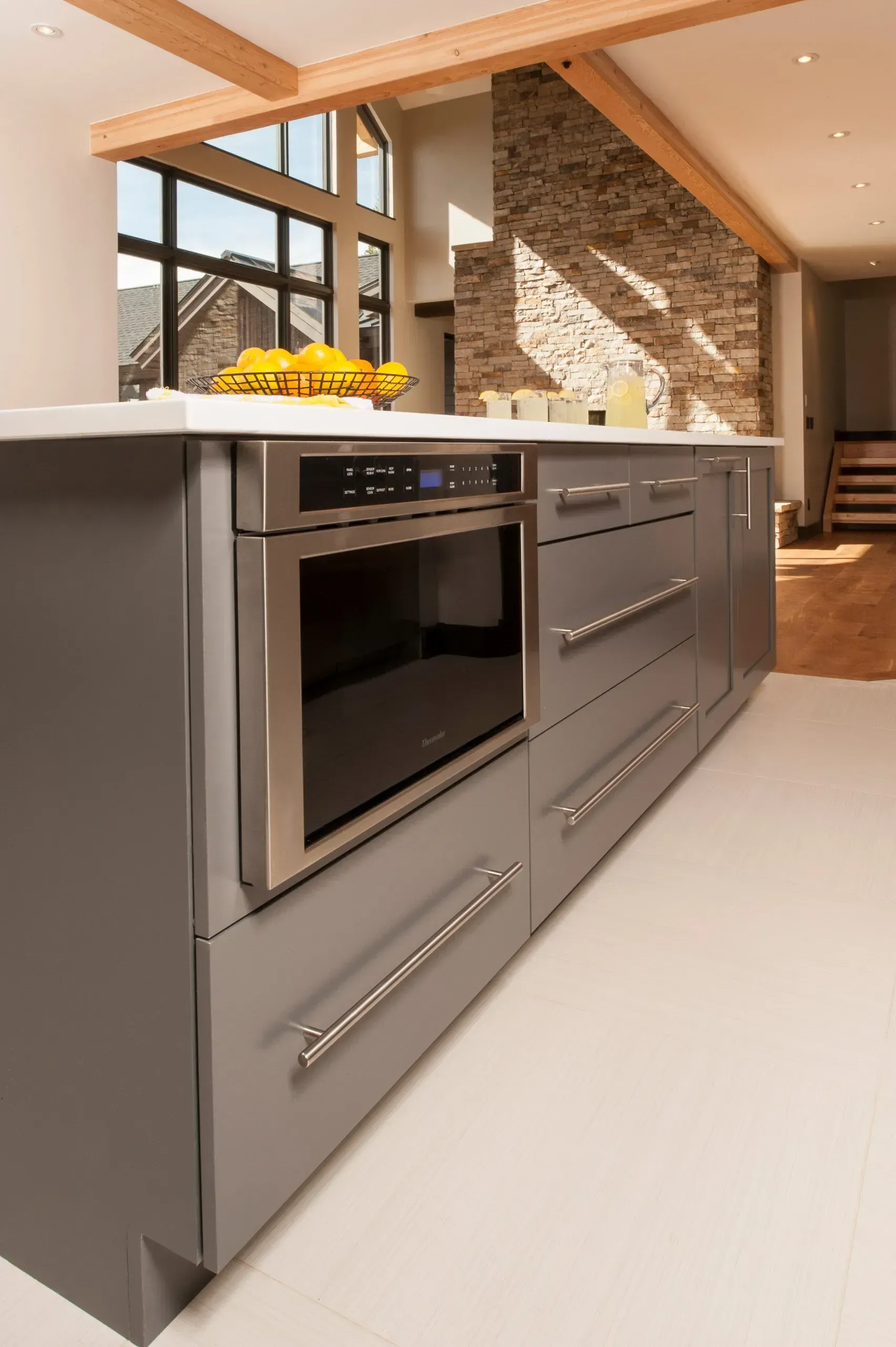 Gray kitchen island with built-in microwave, drawers, and polished chrome handles, against a stone wall and large window.