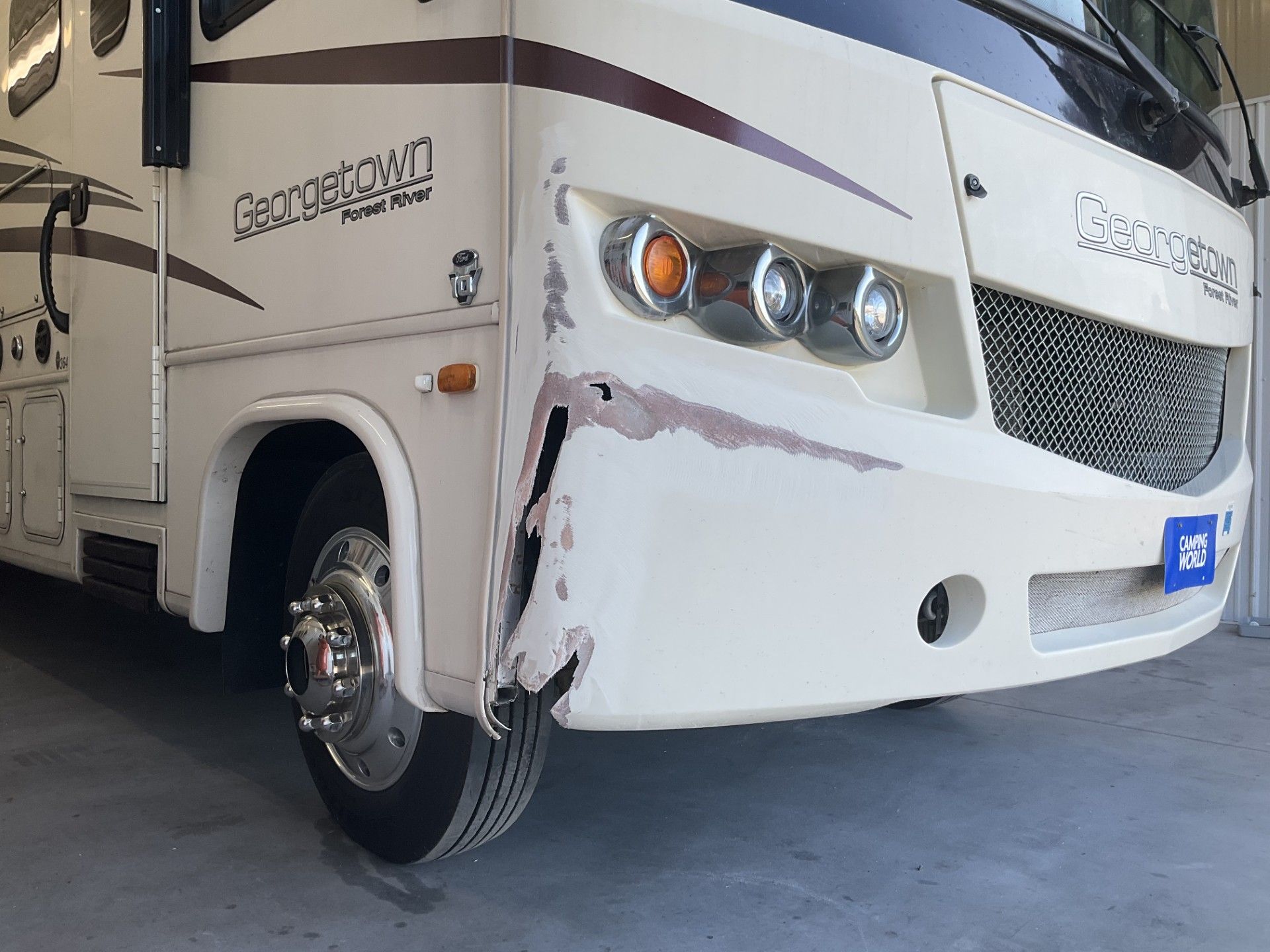 Damaged cream-colored RV front bumper. Shows a dent, scrapes, and exposed metal.