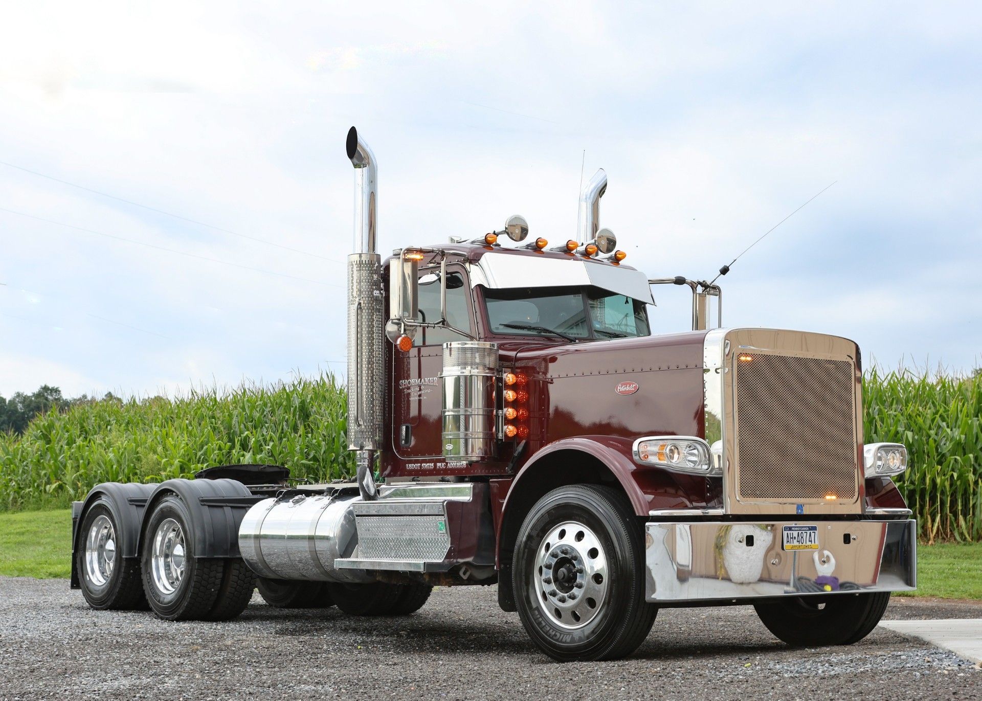 Maroon semi truck parked on a gravel road with green field and cloudy sky
