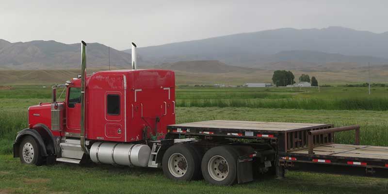 A red semi truck is parked in a grassy field with mountains in the background