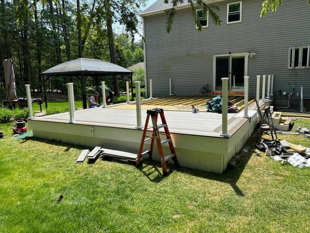 Deck construction in backyard with green grass, house, gazebo, and ladder present.