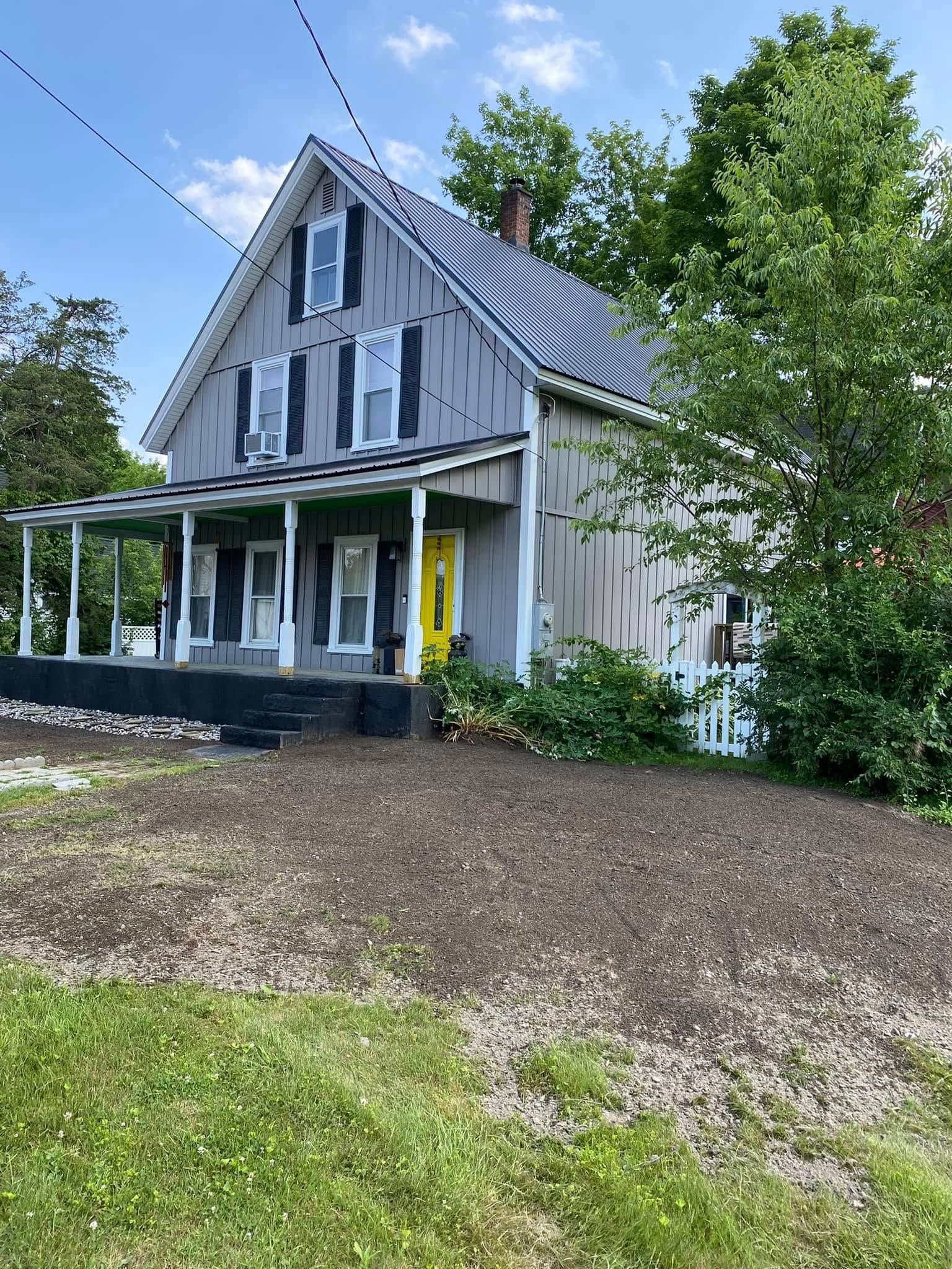 Two-story grey house with yellow door, black shutters, and a porch. Overgrown yard.