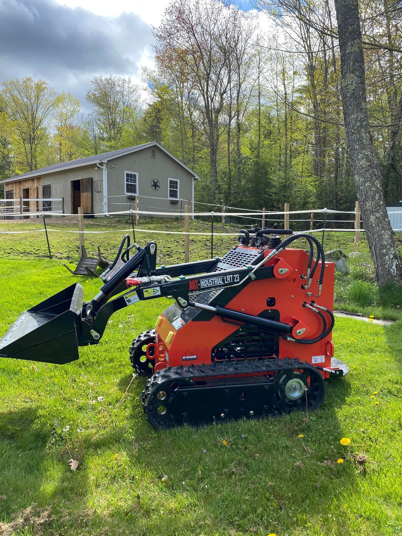 Orange mini skid steer loader on grass in front of a small building and trees.
