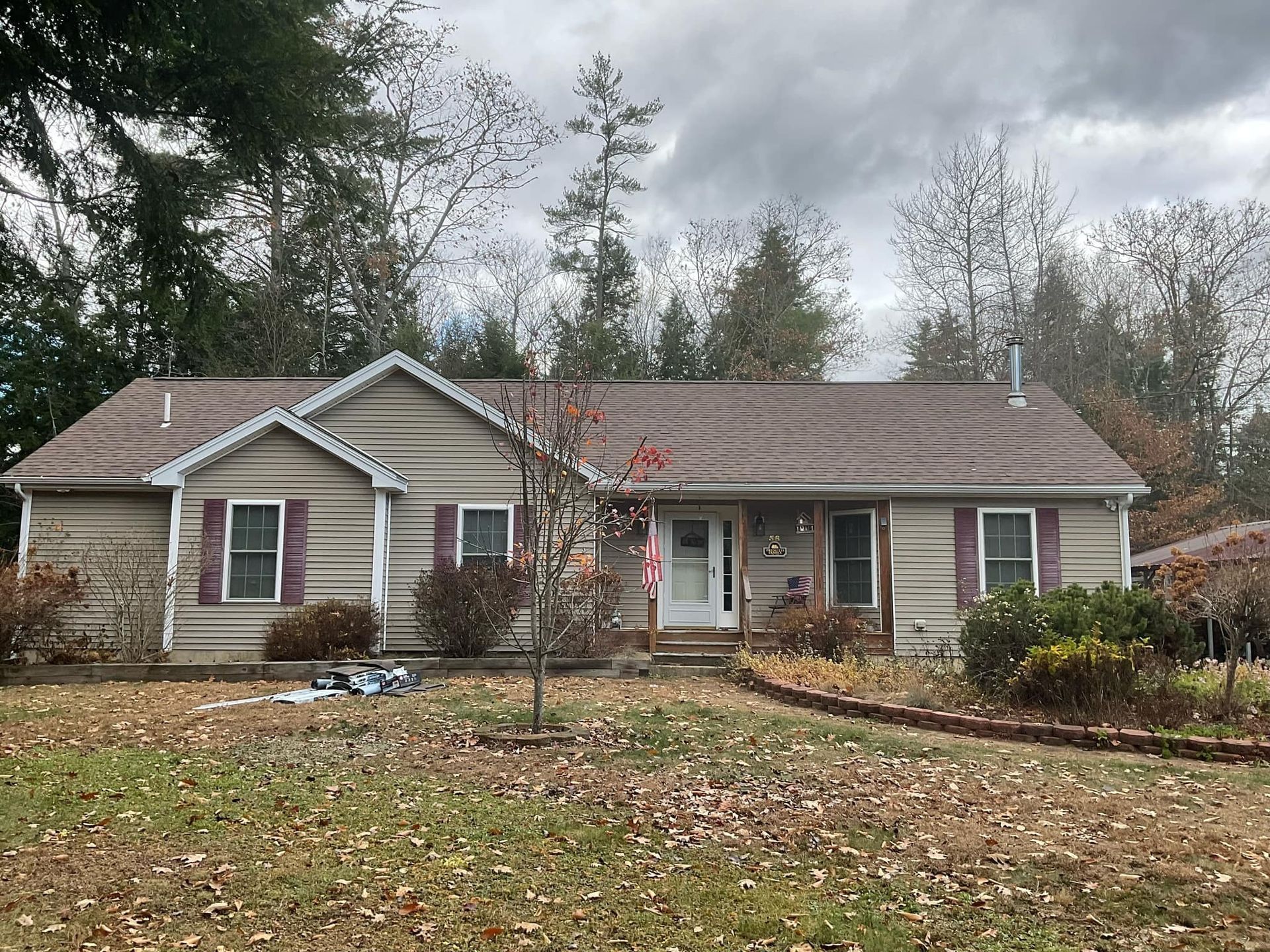 A one-story house with beige siding and maroon shutters, in a yard with fallen leaves, surrounded by trees.