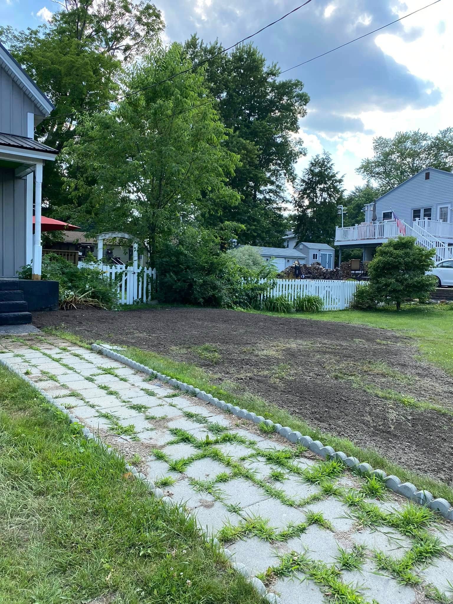 A paved path leading to a brown dirt garden bed, a house on the left, and a white fence in the background.