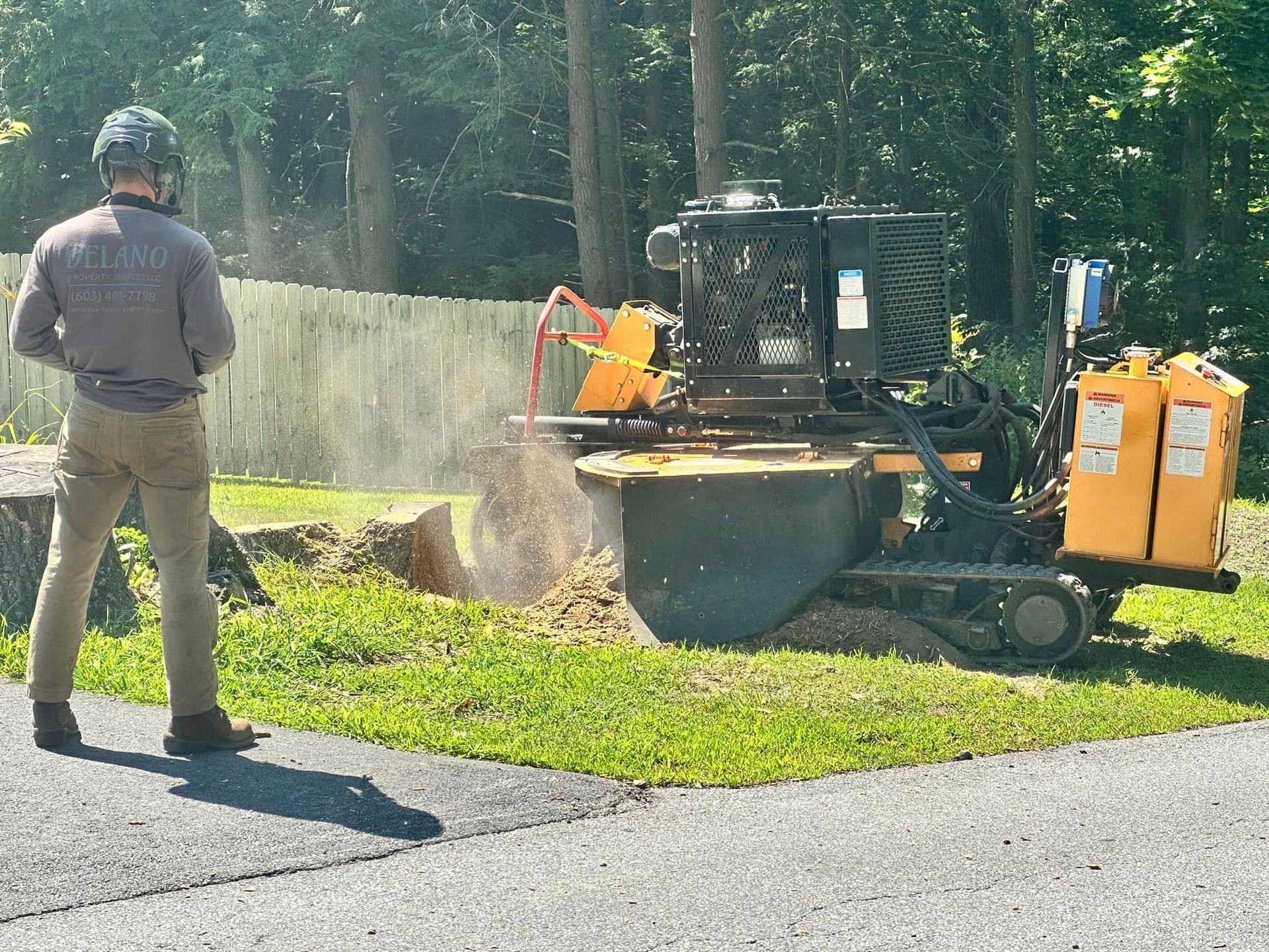 Man operating a yellow stump grinder on a tree stump, creating wood chips on grass.