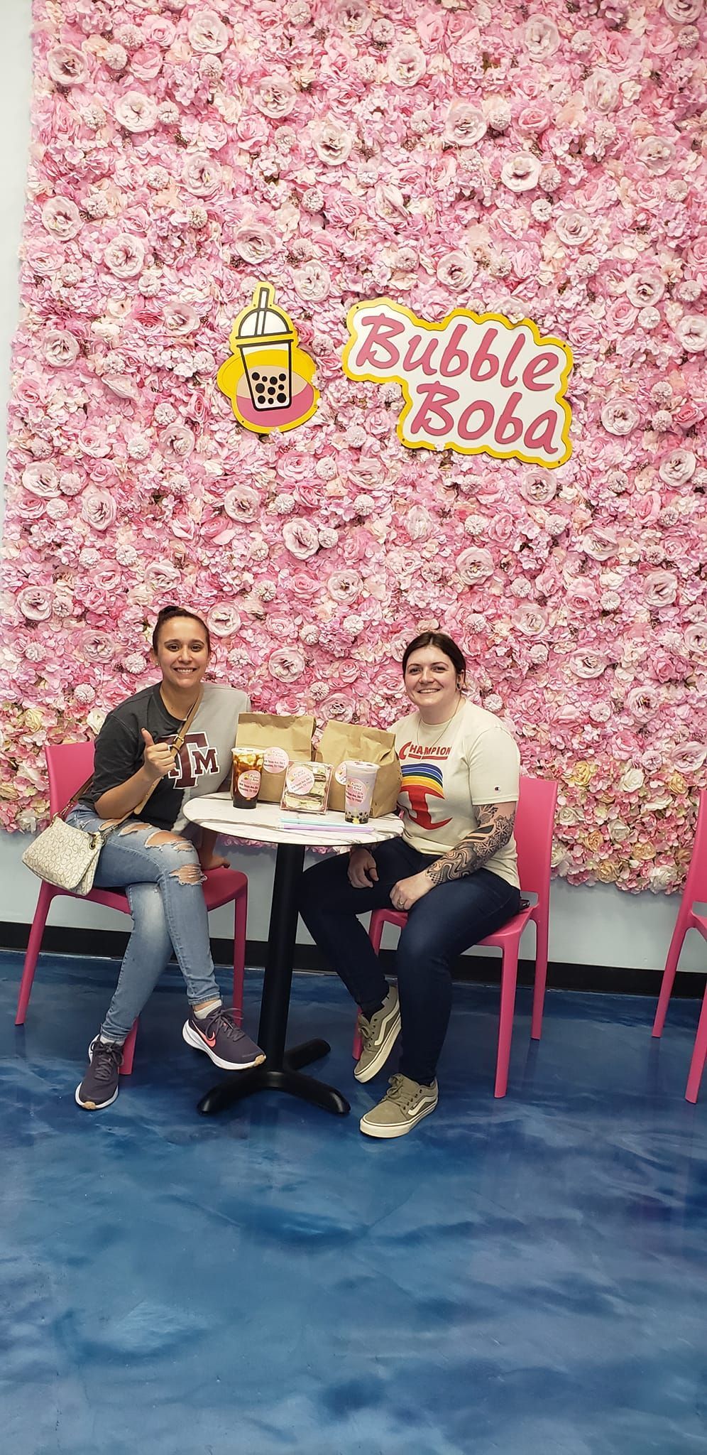 Two women are sitting at a table in front of a pink flower wall.