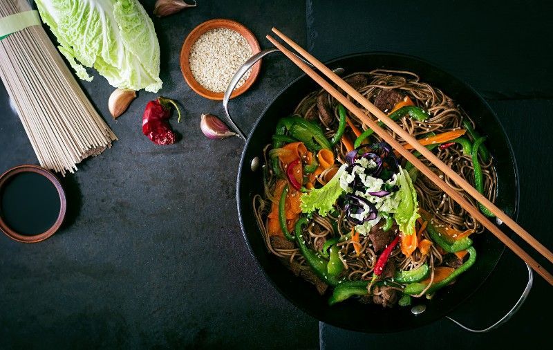 A wok filled with noodles and vegetables with chopsticks on a table.