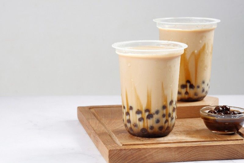 Two cups of bubble tea are sitting on a wooden cutting board.