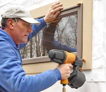 Man installing window frame with a nail gun; tan frame, blue jacket, outside.
