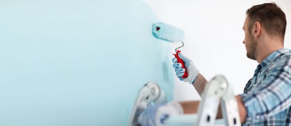 Man painting a wall blue with a paint roller. He wears gloves and a plaid shirt.