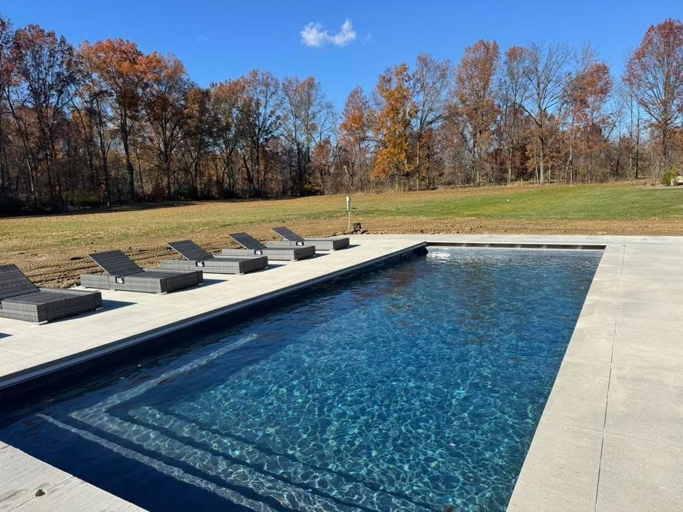 Pool with dark blue water and lounge chairs on a concrete deck, surrounded by a lawn and trees.