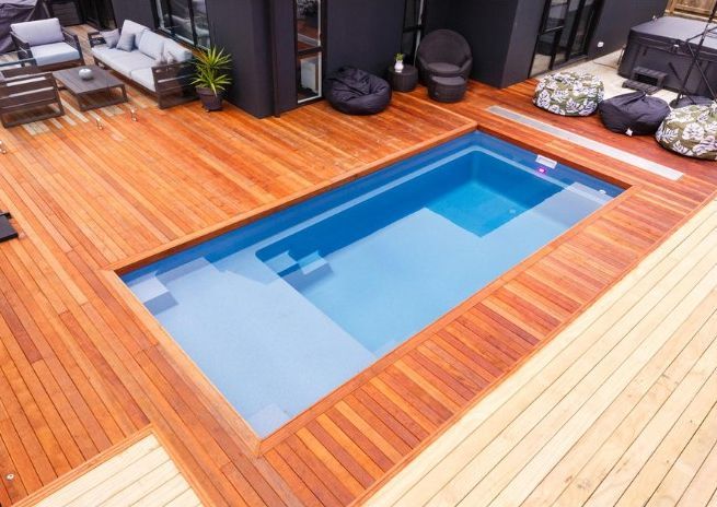 Wooden deck surrounds a rectangular blue swimming pool. Outdoor seating and dark exterior of a building visible.
