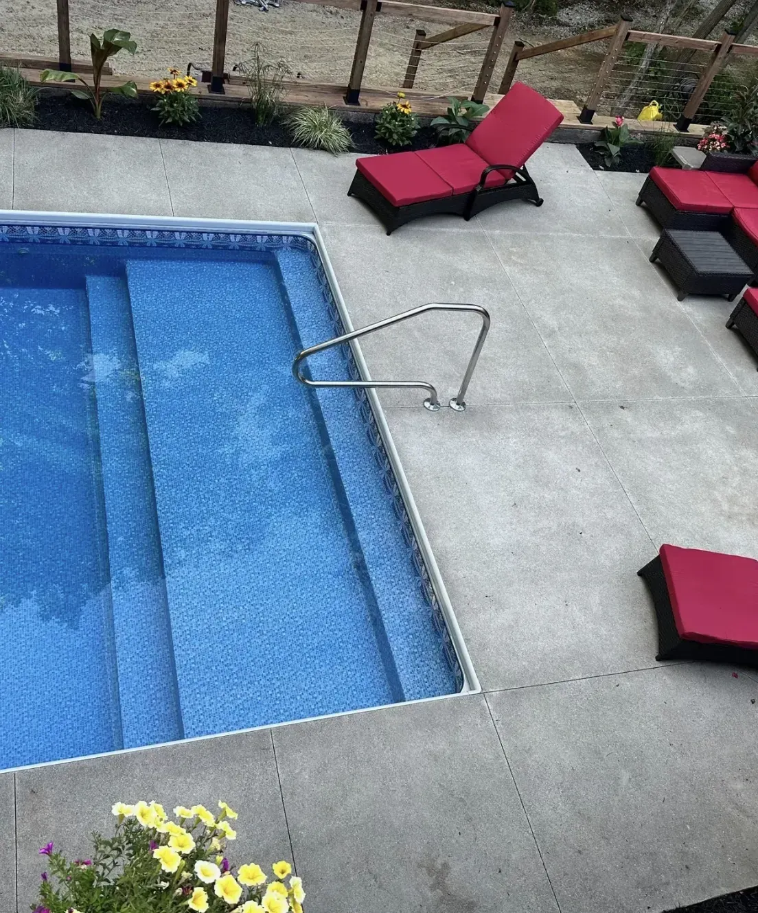 Swimming pool with built-in steps and handrail, surrounded by concrete patio and red lounge furniture.