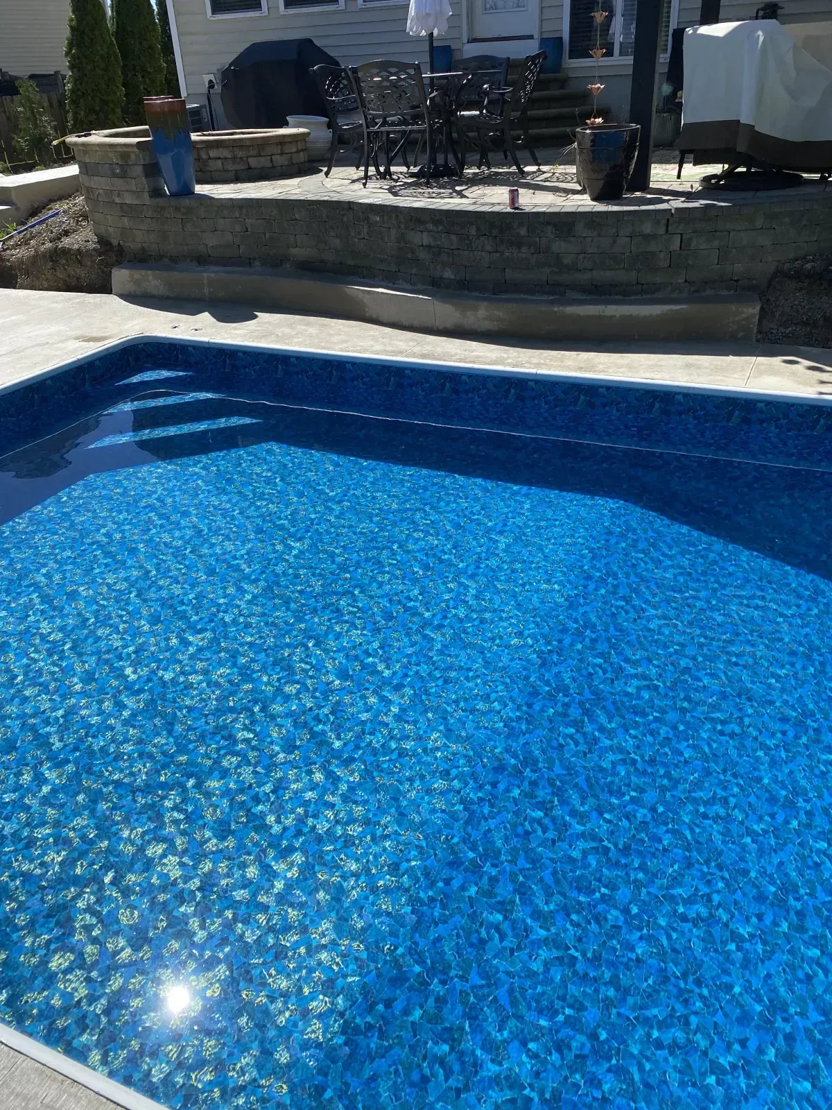 A blue swimming pool with steps, surrounded by a patio and stone wall.