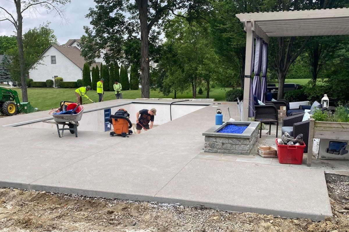Concrete patio being built near a pool; workers, tools, and pergola present.