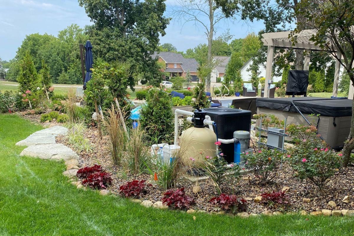 Backyard landscape with a pool filter, hot tub, and various plants. Sunny day with trees in the background.