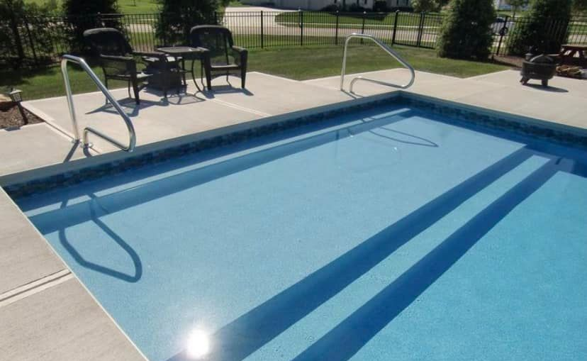 Outdoor swimming pool with built-in steps and handrails, bordered by concrete. Two chairs and a small table are nearby.