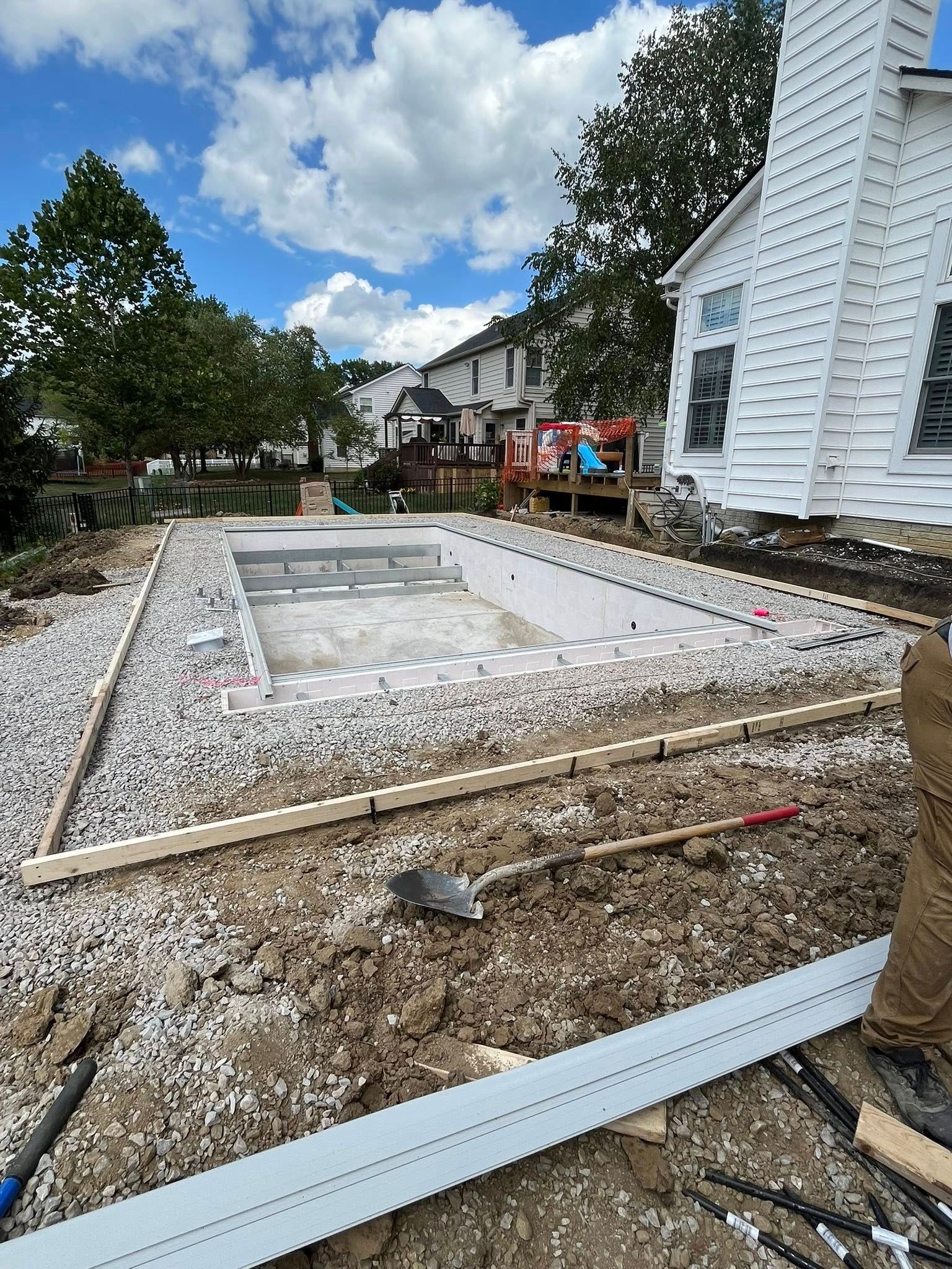 Pool construction site with gravel, wooden forms, and a house. Blue sky overhead.