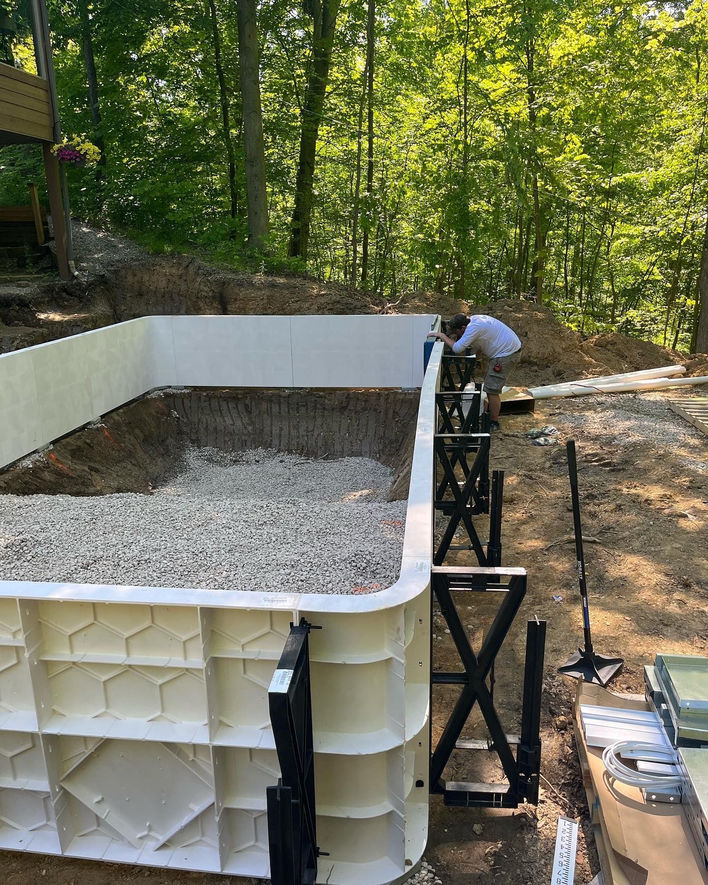 Pool construction: Man working on a partially built, white-walled pool with gravel base in a wooded area.