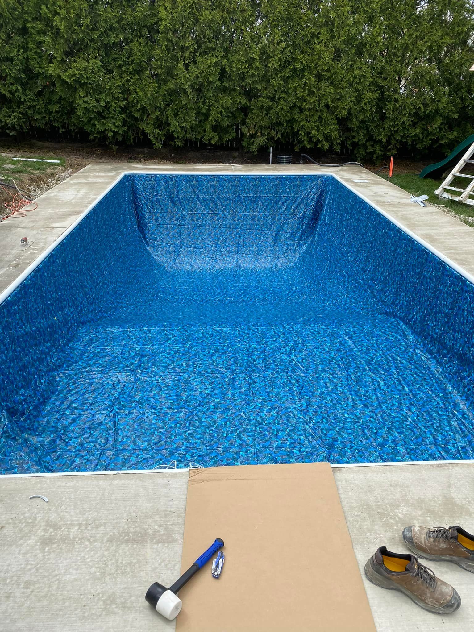 Empty blue tiled swimming pool, construction materials and work boots on concrete surround.