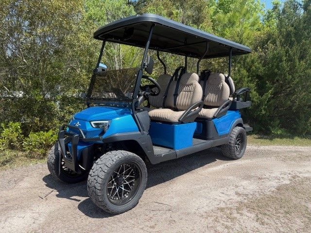 Blue six-seat golf cart parked on a dirt path beside green trees.