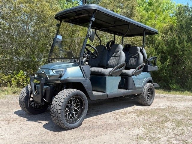 Green six-seat golf cart with roof parked on a dirt path by trees.