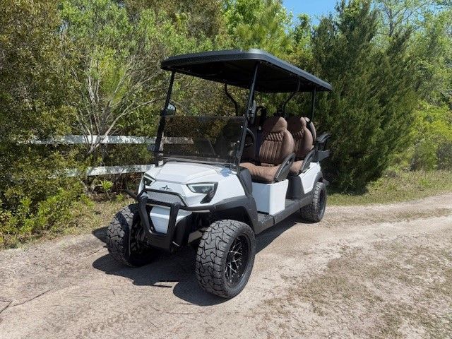 White six-seat golf cart with a canopy on a dirt path beside green shrubs and trees.