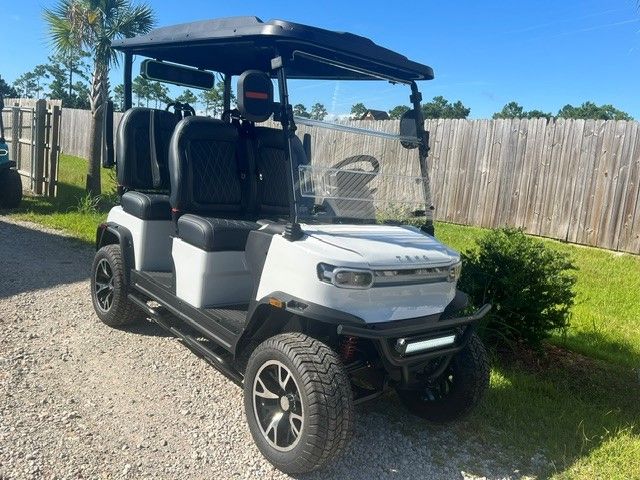 White golf cart parked on gravel beside a wooden fence and shrubs under a blue sky