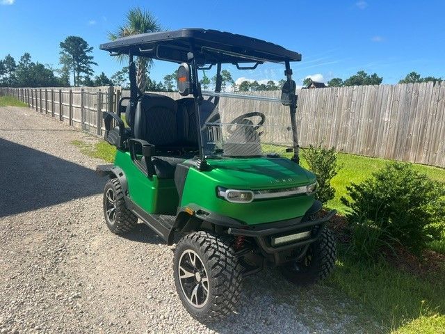 Green utility ATV parked on a driveway beside a fence and shrubs under a sunny sky