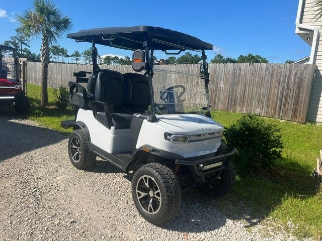 White golf cart with black roof parked on a gravel driveway beside a wooden fence and greenery.