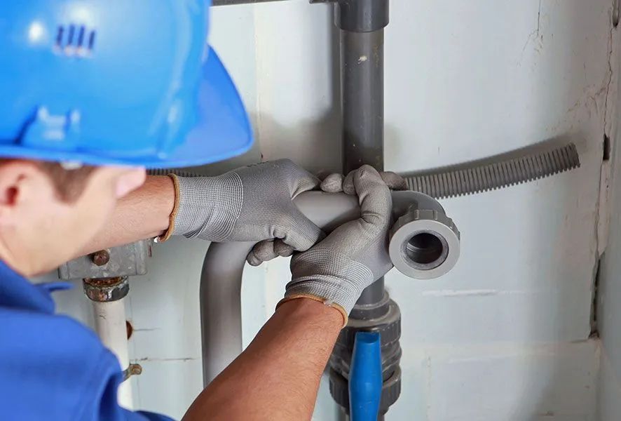Plumber in blue uniform and hard hat connecting pipes in a utility room.