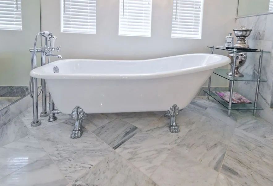 White clawfoot bathtub in marble-tiled bathroom with window blinds.
