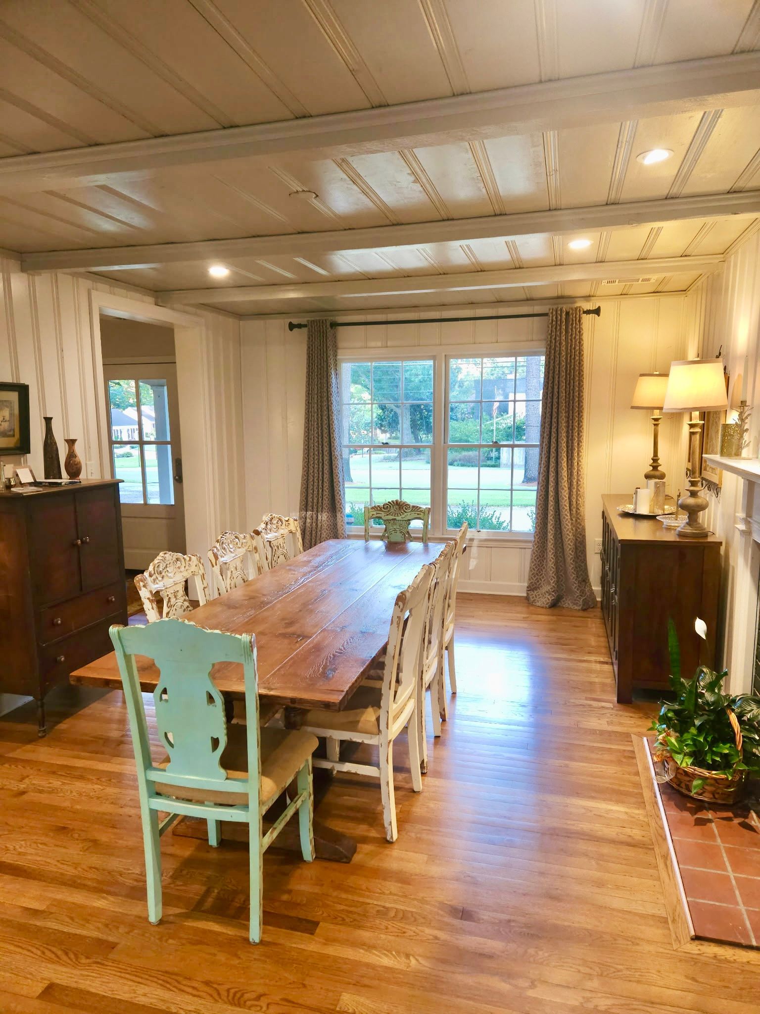 A dining room with a long wooden table and chairs. Wood floors and ceiling beams. Window with curtains in the back.
