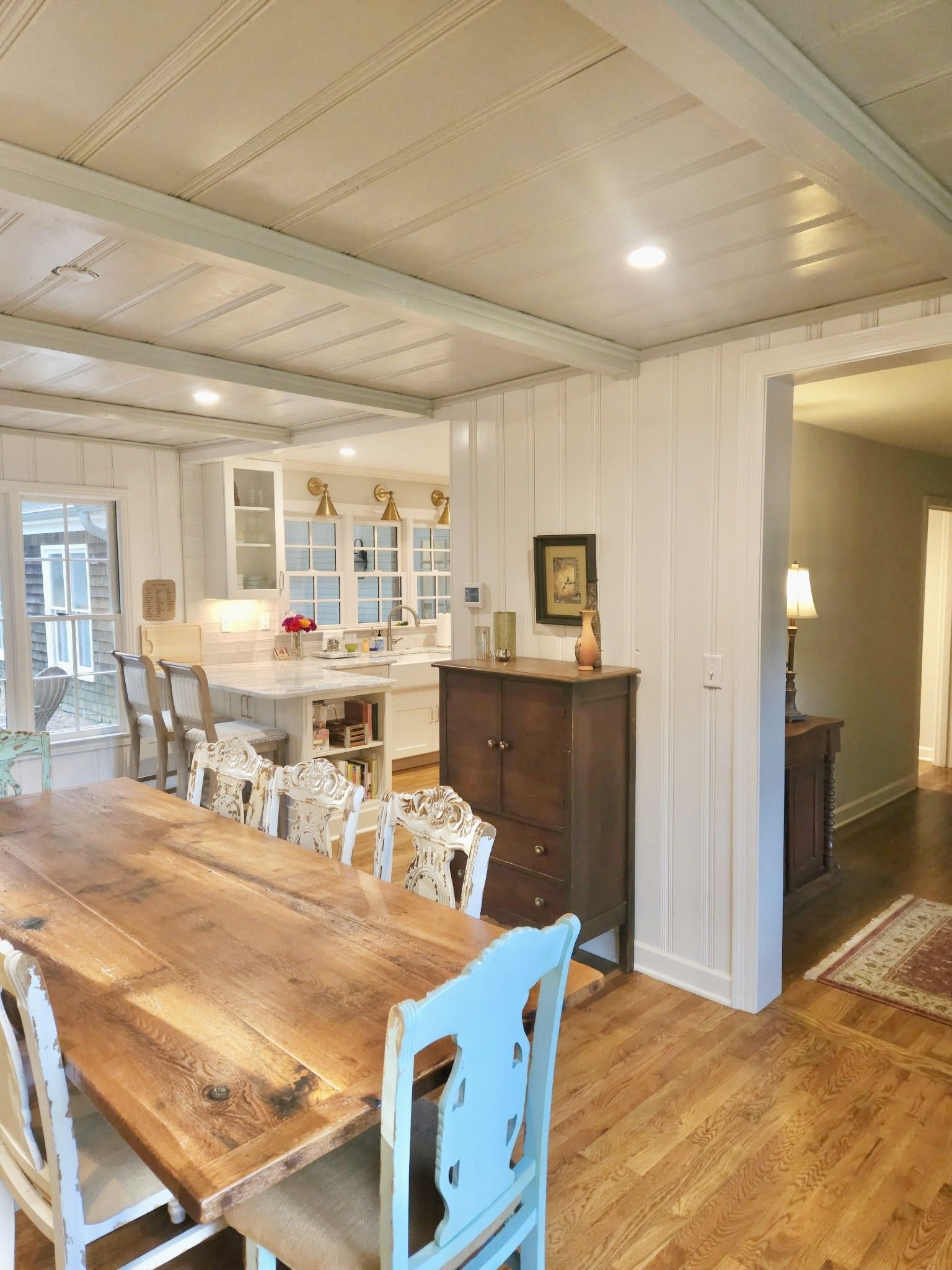 A dining room with a long wooden table, white chairs, and a view into the kitchen.