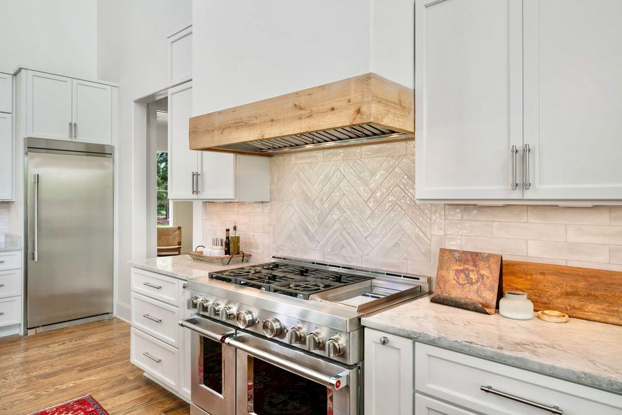 Modern white kitchen with stainless steel appliances, marble countertops, and wooden range hood.