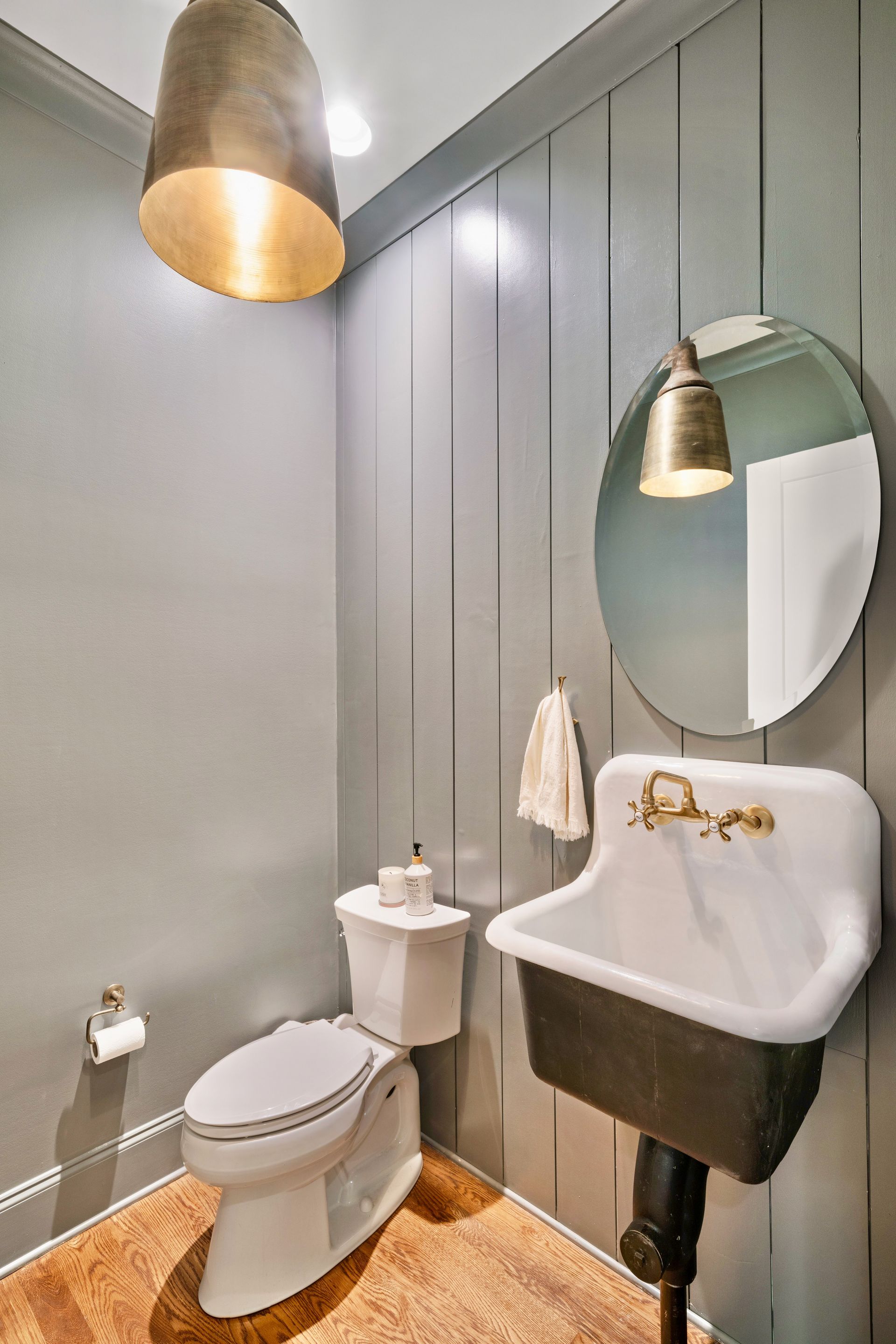 Small, gray-walled bathroom with toilet, sink, mirror, and two bell-shaped light fixtures. Cork flooring.