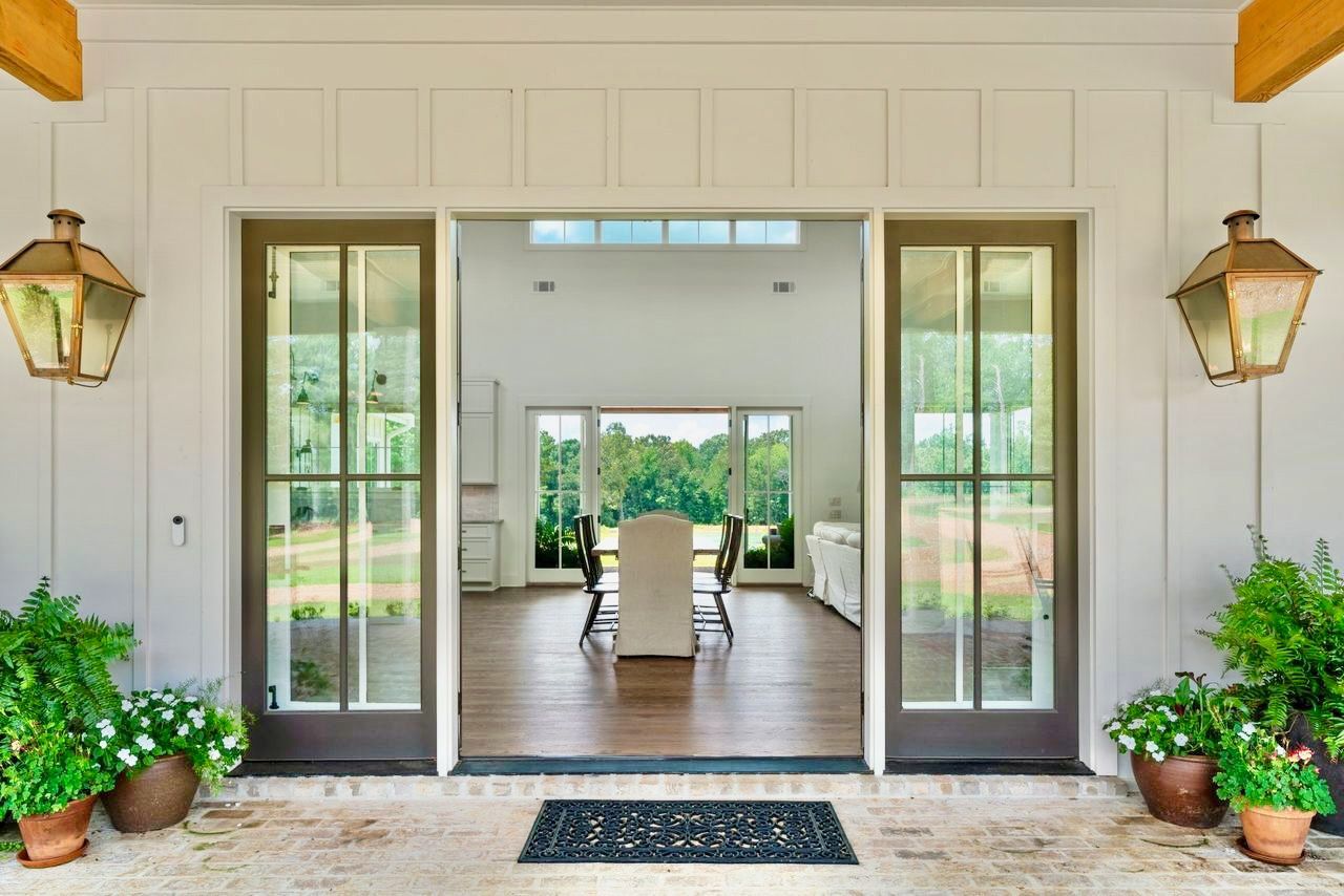 Open double doors reveal a dining room with a table, surrounded by windows and lush greenery.