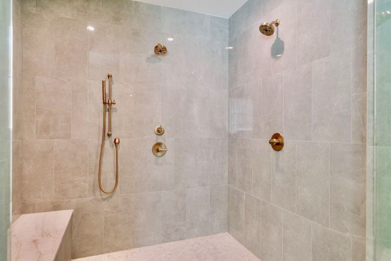 Modern shower with gold fixtures and light gray tiled walls, a bench, and glass door.