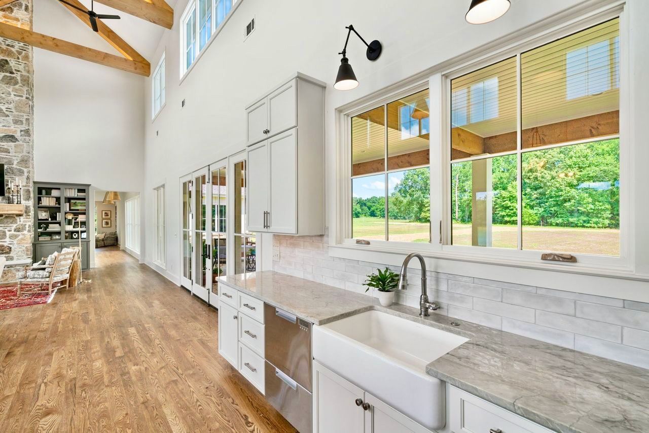 Spacious kitchen with wood floors, white cabinets, farmhouse sink, large windows overlooking a green yard.
